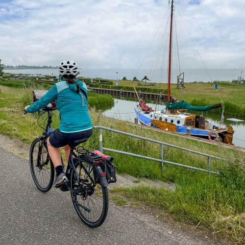 Female cyclist in turquoise jacket riding on paved dike path along IJsselmeer with traditional sailboat moored in canal and green marshland.