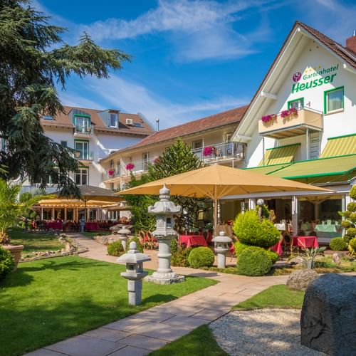 Gartenhotel Heusser in Bad Dürkheim mit weißer Fassade, grünen Markisen, Terrasse mit gelben Sonnenschirmen und Garten mit asiatischen Steinlaternen.