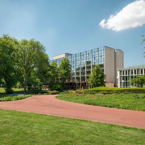 Modern Sheraton Essen hotel building with glass facade surrounded by green parkland, trees and a red brick pathway under blue sky.
