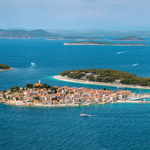 Luftaufnahme der Halbinsel Primošten mit terrakottafarbenen Dächern, Kirchturm und türkisfarbenem Adriatischem Meer. Inseln und Berge im Hintergrund.