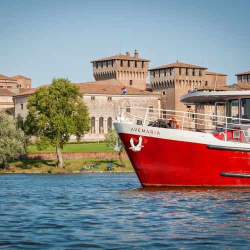Rot-weißes Passagierschiff MS Ave Maria auf blauem Wasser mit historischen Festungsmauern und Türmen in Mantua, Italien unter klarem Himmel.