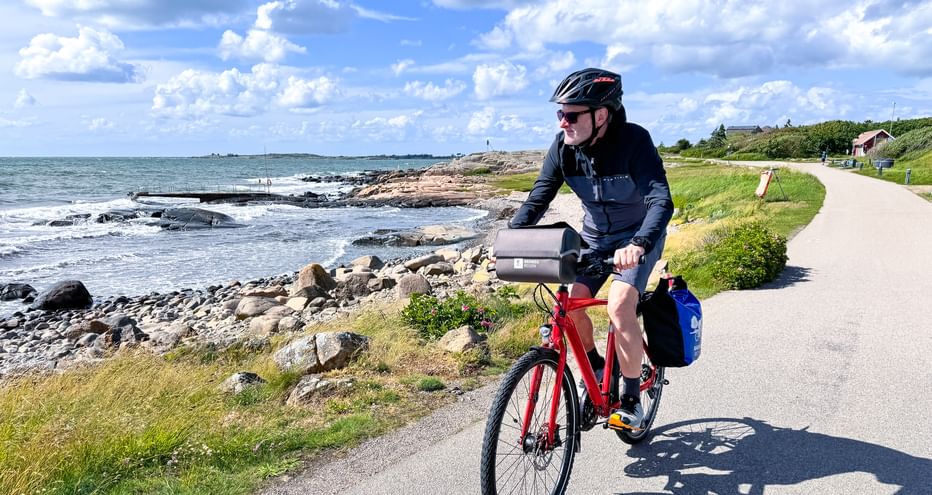 Cyclist with helmet riding red bike on coastal path along Swedish west coast near Varberg, with rocky shoreline and blue sea under cloudy sky.