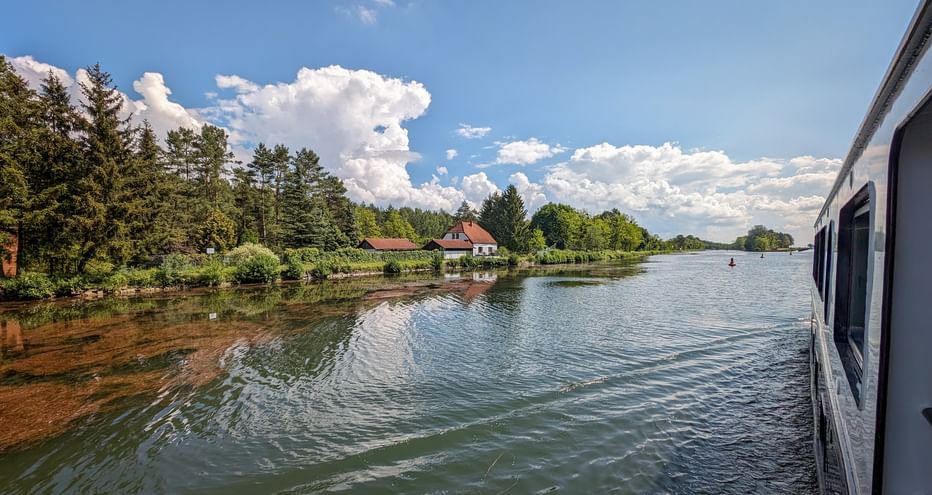 View from MS Princess ship deck showing a calm waterway with houses and trees along the shore under a blue sky with white clouds.