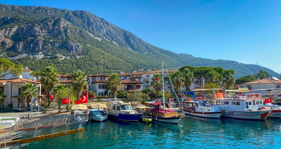 Hafen von Akyaka mit bunten Booten unter türkischen Flaggen im türkisfarbenen Wasser. Weiße Häuser mit Palmen und grüner Berg unter blauem Himmel.