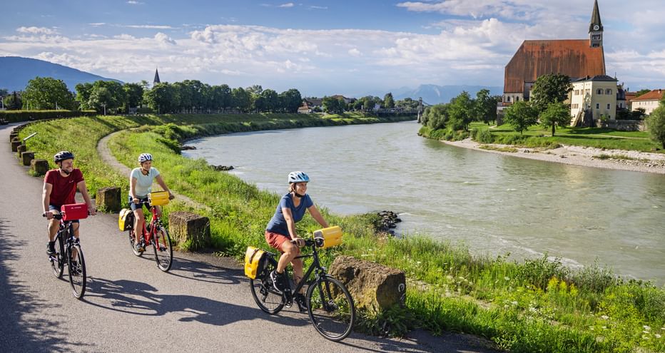 Three cyclists with yellow panniers riding along the Salzach River path near Laufen. The baroque Stiftskirche church is visible across the river.
