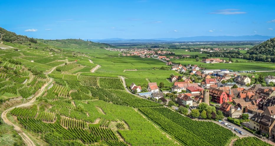 Luftaufnahme des Dorfes Ammerschwihr im Südelsass, umgeben von terrassierten Weinbergen unter blauem Himmel mit fernen Bergen.