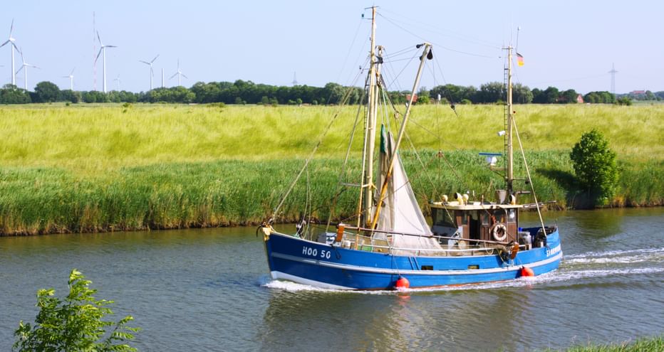 Ein kleines Fischerboot auf einem kleinen Fluss umgeben von Feldern, im Hintergrund Windräder
