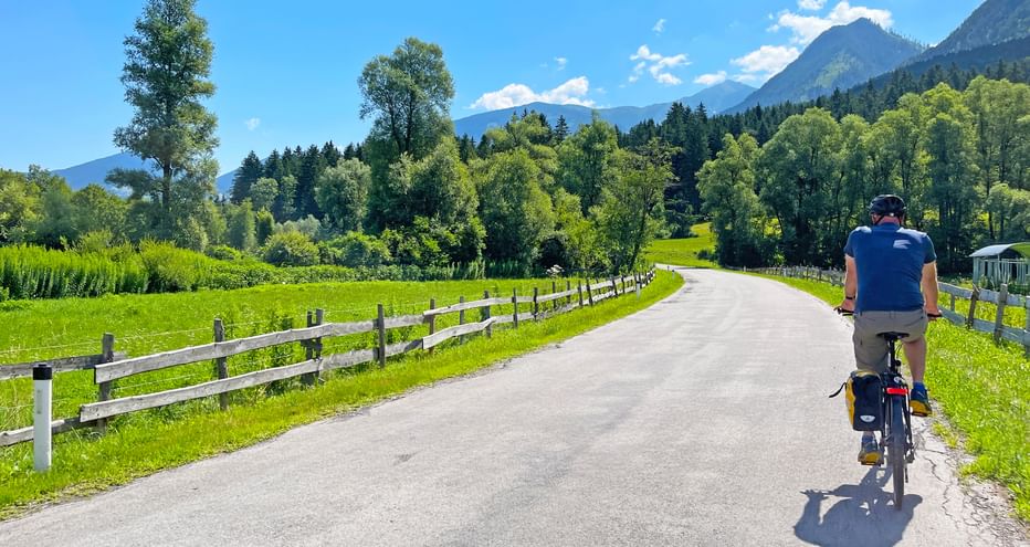 Radfahrer auf asphaltierter Straße, umgeben von Wiesen, Bäumen und Bergen