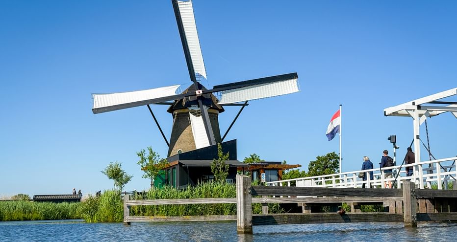 Traditional Dutch windmill with white sails at Kinderdijk beside a canal. A white wooden bridge with Dutch flag and people crosses the waterway.