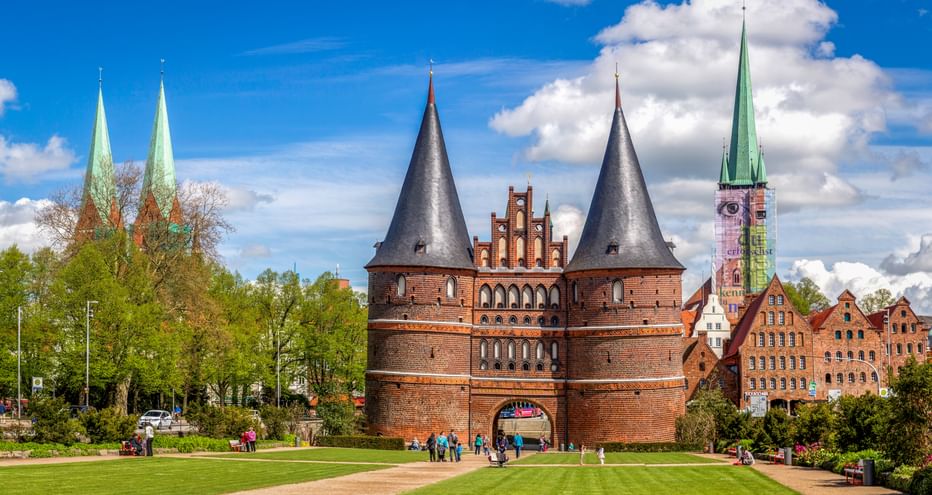 Historic Holstentor gate with twin towers in Lübeck, surrounded by green lawn and trees. Church spires and historic buildings visible in background.