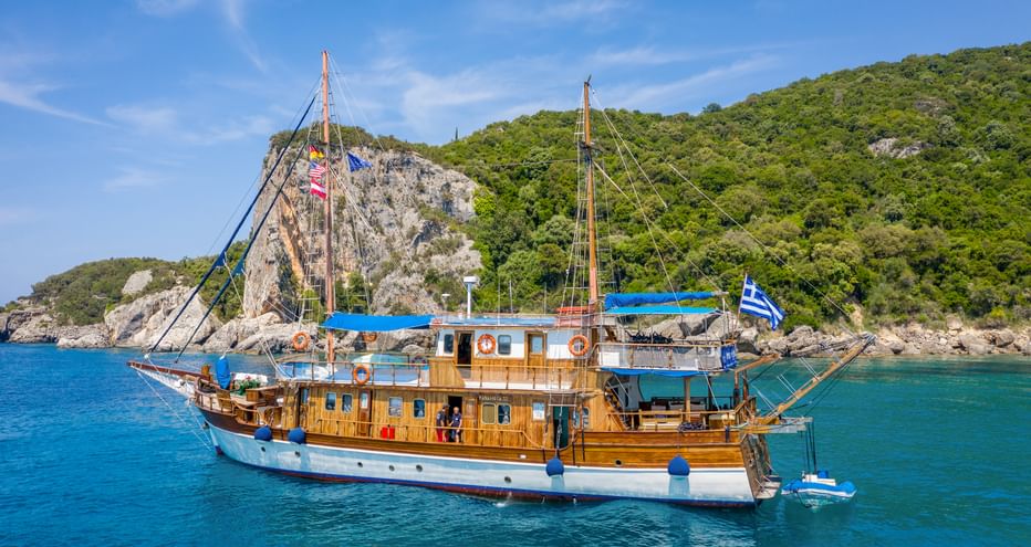 Traditionelles Holzsegelschiff MS Panagiota mit blauem Sonnensegel in türkisfarbener Bucht. Grüne bewaldete Hügel erheben sich aus dem Wasser.