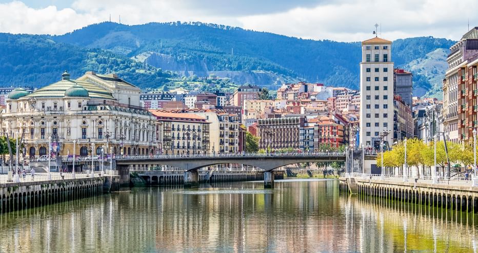 River view of Karlsbad with historic buildings, a bridge, and forested mountains in the background under a partly cloudy sky.