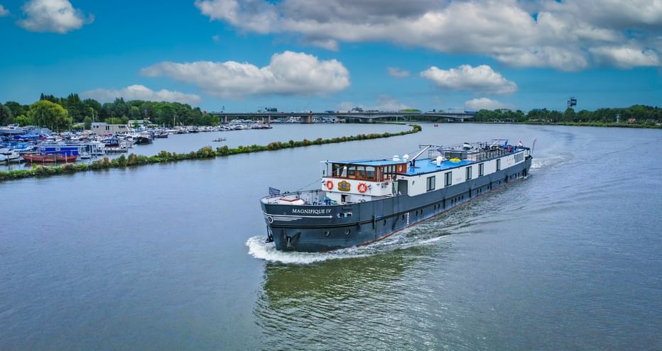 Flusskreuzfahrtschiff MS Magnifique II fährt auf breiter Wasserstraße unter blauem Himmel mit weißen Wolken. Marina und grüne Bäume am linken Ufer.