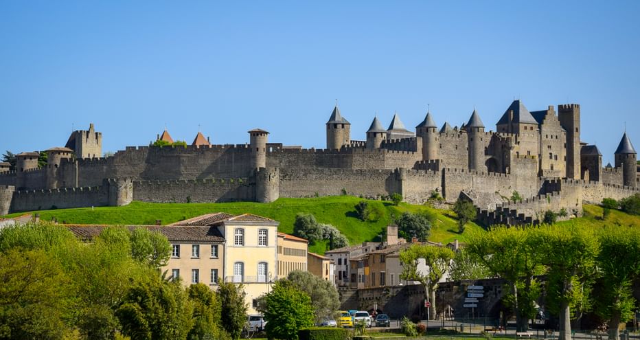 Medieval fortress of Carcassonne with stone walls and towers on a hill, surrounded by green grass and trees. Residential buildings in foreground.