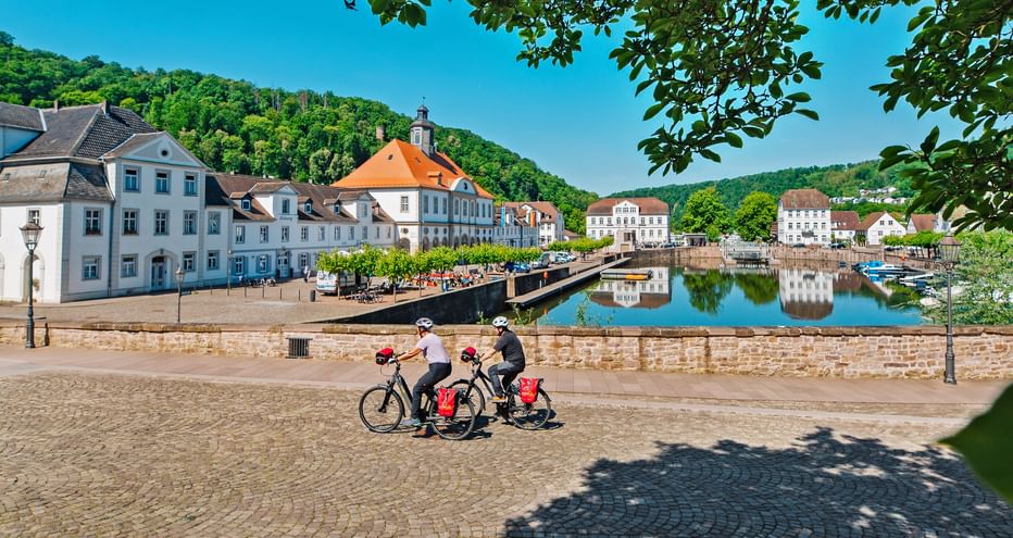 Zwei Radfahrer mit roten Taschen fahren am Barockhafen in Bad Karlshafen entlang, mit historischen weißen Gebäuden und einer Brücke im ruhigen Wasser.