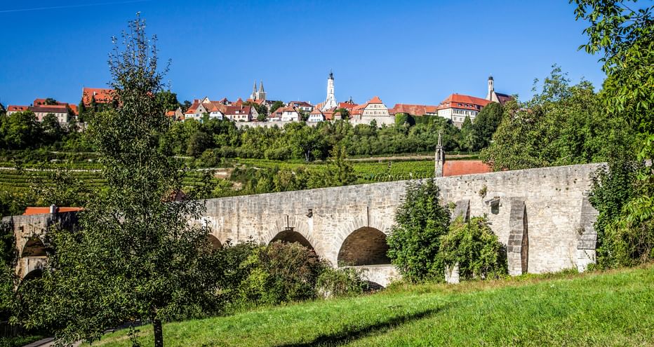 Historische Steinbrücke mit Bögen im Vordergrund, mittelalterliche Stadt Rothenburg ob der Tauber mit rotgedeckten Gebäuden und Kirchtürmen auf Hügel.