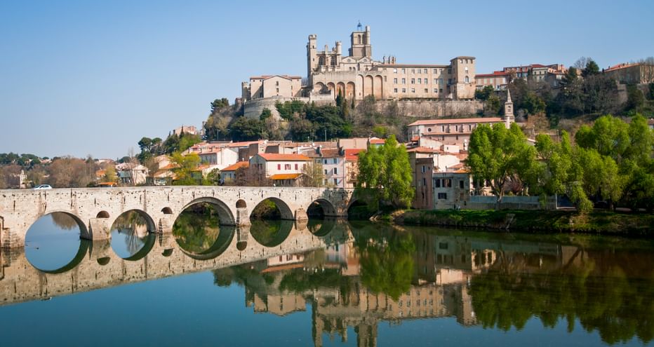 Steinbrücke mit mehreren Bögen über einem ruhigen Fluss in Béziers, mit der Kathedrale Saint-Nazaire auf dem Hügel und Spiegelungen im Wasser.
