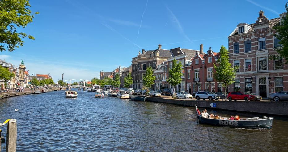 Spaarne canal in Haarlem with traditional Dutch houses lining the waterway. Boats moored along the canal, trees, and blue sky visible.