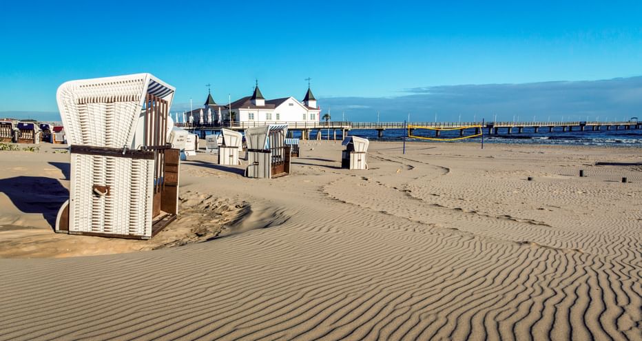Sandstrand mit traditionellen Strandkörben und der Seebrücke Ahlbeck, die sich in die Ostsee auf Usedom erstreckt, unter klarem blauen Himmel.