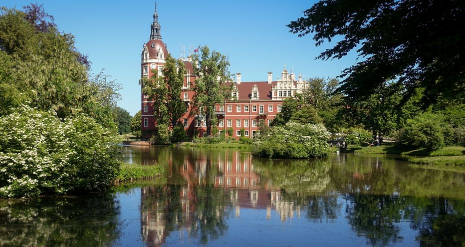 Rotes Backsteinschloss Muskau mit Uhrenturm spiegelt sich im See, umgeben von grünen Bäumen im Fürst-Pückler-Park unter blauem Himmel.
