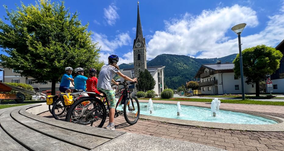 Family of four cycling past a fountain in Maishofen village square, with a church spire and mountains in the background under blue sky.
