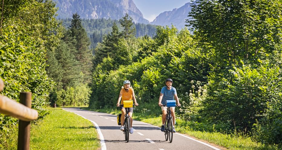 Two cyclists riding on paved path through Kanaltal valley near Tarvis, surrounded by green forest with mountain peaks in background.