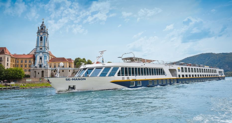 White river cruise ship MS Manon sailing on the Danube near Dürnstein with blue baroque church tower and hills in background.