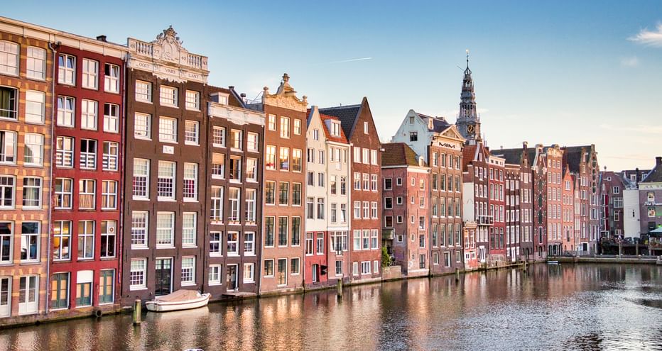 Reihe bunter historischer Grachten­häuser entlang Amsterdams Damrak mit Kirchturm im Hintergrund unter blauem Himmel.