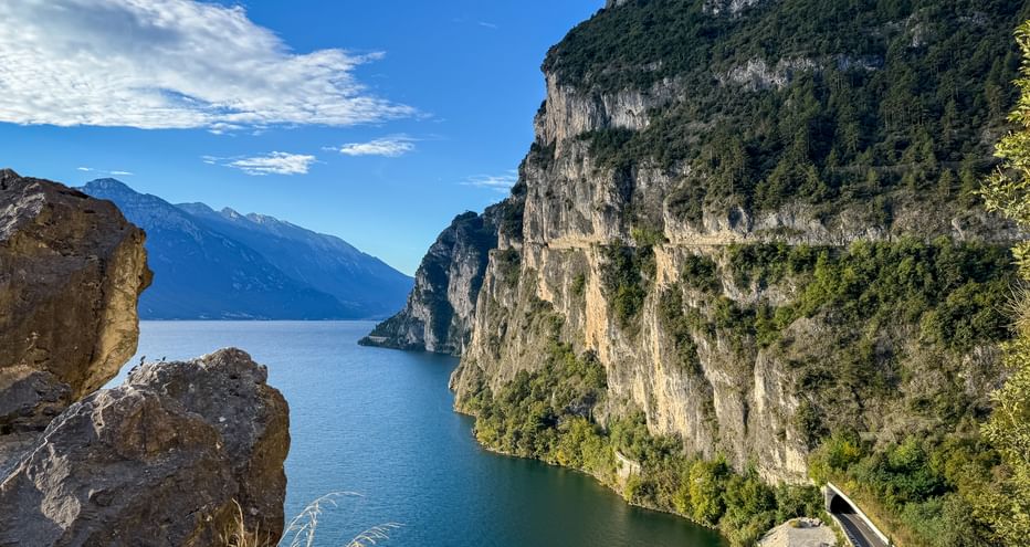 Panoramic view from Strada del Ponale showing steep cliffs, blue Lake Garda waters, and distant mountains under a clear sky.