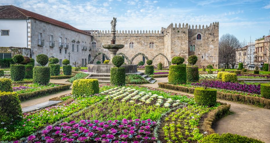 Jardim de Santa Barbara in Braga with colorful flower beds, sculpted hedges, and a fountain. Medieval stone buildings and walls in background.