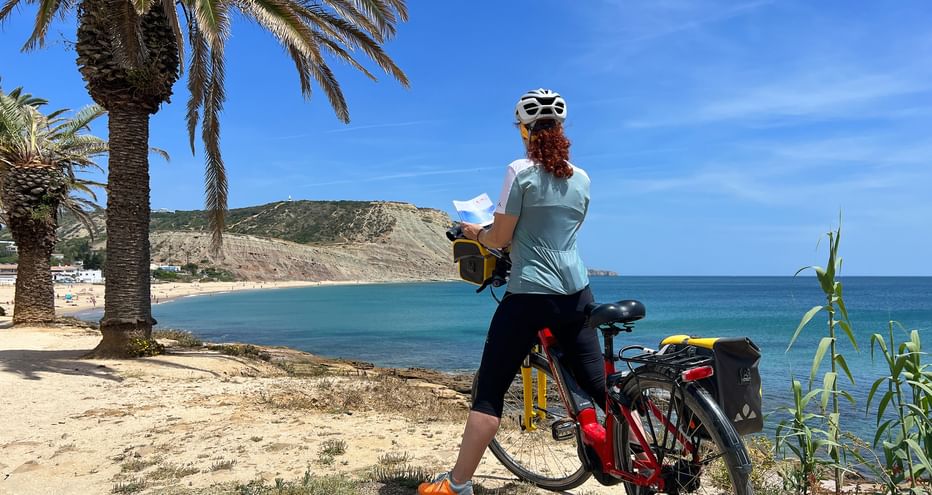 Radfahrerin mit Helm und Karte steht mit Tourenrad bei Palmen an einer Sandbucht mit türkisem Wasser und Hügeln.