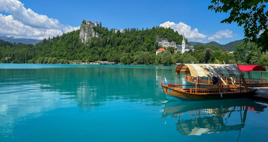 Wooden boat on turquoise Lake Bled with forested hill, Bled Castle, and white church tower. Mountains and blue sky with clouds in background.