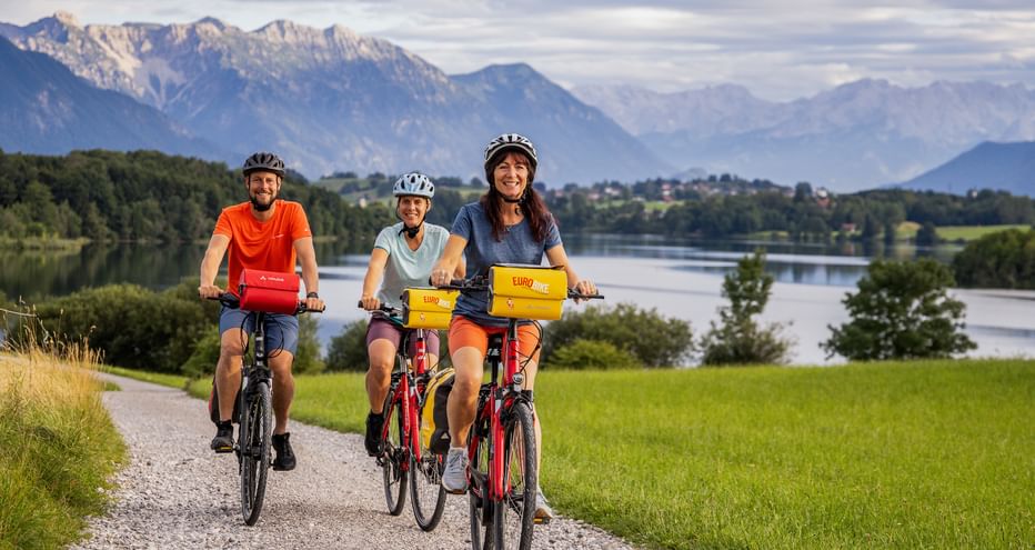 Drei Radfahrer auf einem Schotterweg am Riegsee in Bayern, mit grünen Wiesen und dramatischen Alpenbergen im Hintergrund.