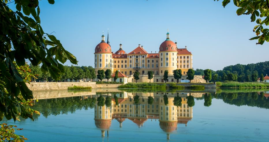 Moritzburg Castle with yellow facade and red domes perfectly reflected in calm water, framed by green foliage under blue sky near Dresden.