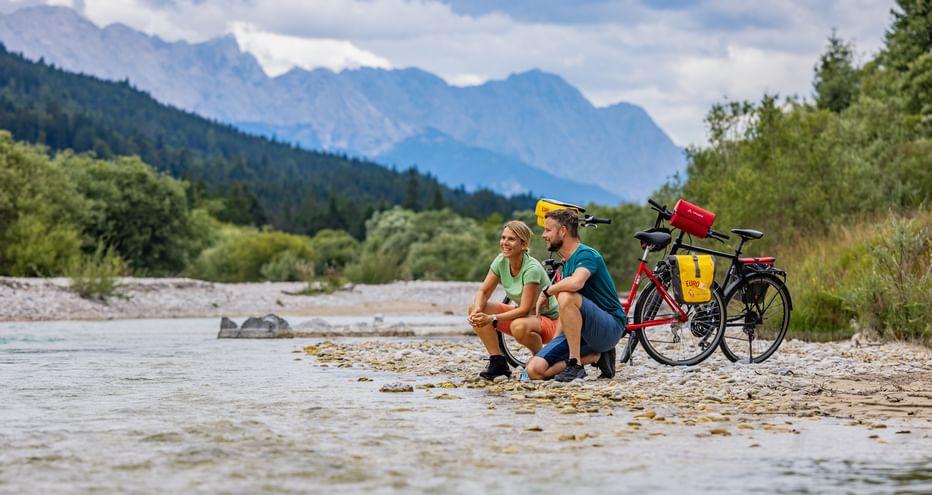 Two cyclists sitting on a pebble beach by the Isar River with touring bikes. Mountains and forest visible in the background under cloudy sky.