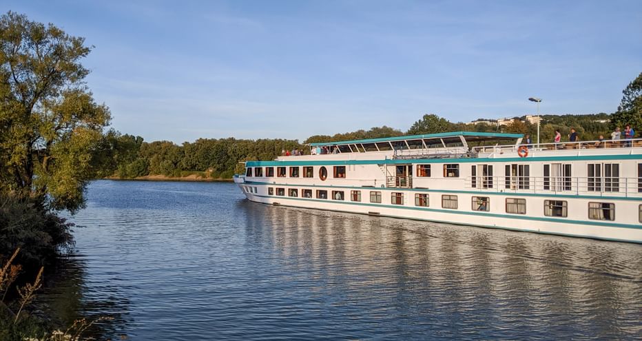 MS Switzerland river cruise ship with white hull and turquoise trim sailing on calm river, surrounded by green trees under blue sky.