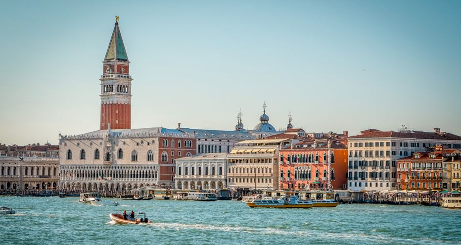 Venedigs Uferpromenade mit Markusturm und bunten historischen Gebäuden. Boote fahren durch die türkisfarbene Lagune unter blauem Himmel.