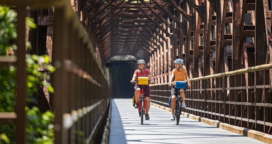 Two cyclists riding through a historic iron railway bridge with wooden truss structure in the Kanaltal valley on the Alpe Adria cycle route.