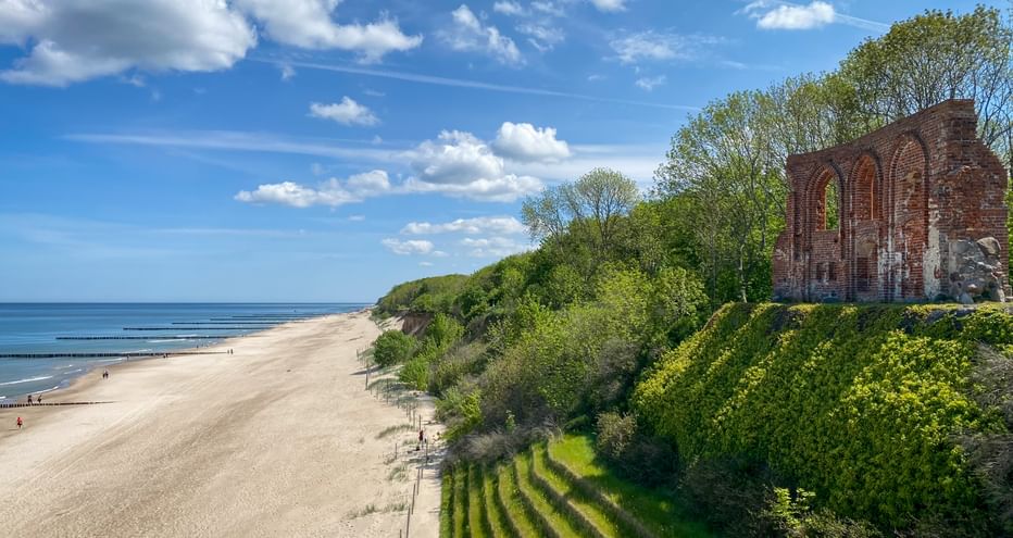 Red brick ruins of St. Nicholas Church on a coastal cliff in Trzęsacz, overlooking a sandy beach and the Baltic Sea under blue sky.