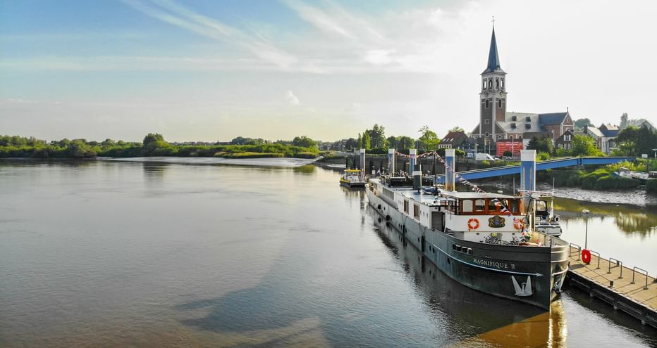 Flusskreuzfahrtschiff MS Magnifique 2 am Holzsteg in Sint-Amands. Eine Kirche mit hohem Turm ist im Hintergrund unter blauem Himmel sichtbar.