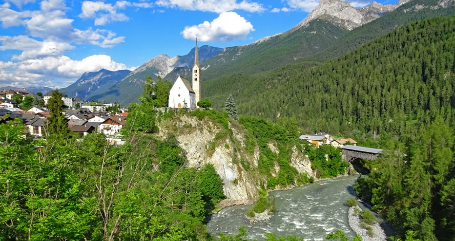Blick auf das Bergdorf Scuol an einem Gebirgsbach, umgeben von Wäldern und Bergen