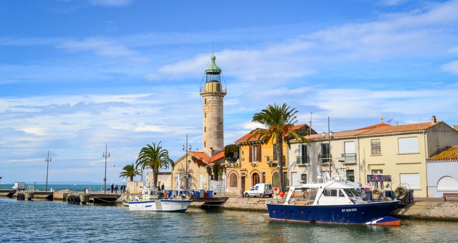 Stone lighthouse at Le Grau-du-Roi harbor with fishing boats, Mediterranean houses, palm trees and blue sky in Camargue, Provence.