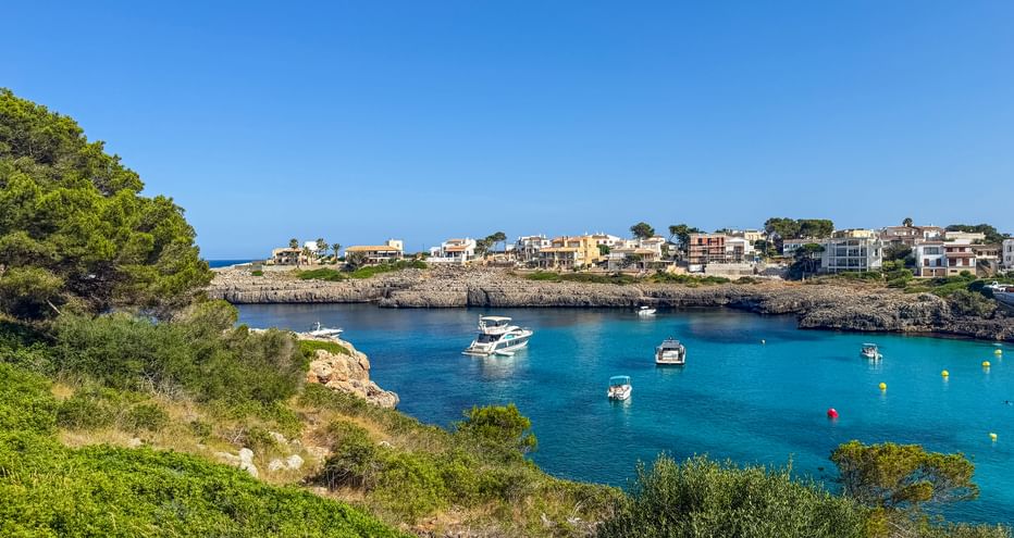 Turquoise bay with boats anchored in calm waters near Porto Colom, Mallorca. Rocky coastline with green vegetation and Mediterranean houses visible.