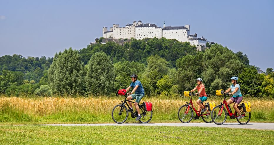 Drei Radfahrer mit gelben Taschen fahren auf einem Weg durch grüne Wiesen mit der Festung Hohensalzburg auf einem bewaldeten Hügel im Hintergrund.