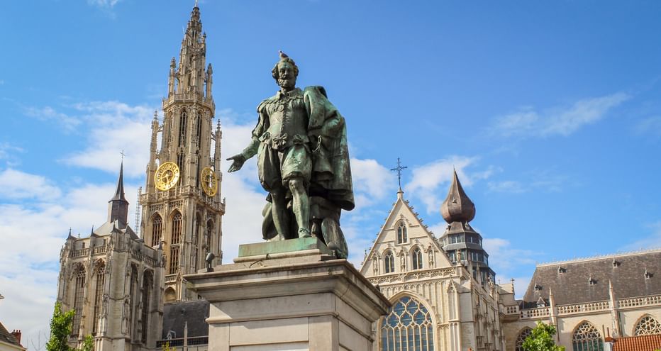 Bronzestatue von Peter Paul Rubens auf einem Sockel vor der Liebfrauenkathedrale in Antwerpen mit ihrem hohen gotischen Turm unter blauem Himmel.