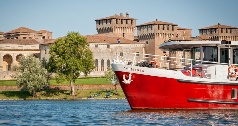 Rot-weißes Passagierschiff Ave Maria auf blauem Wasser vor historischen Festungsmauern und Türmen in Mantua mit grünen Bäumen am Ufer.