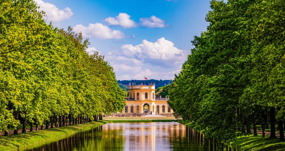 Yellow Orangerie palace in Kassel framed by symmetrical rows of green trees along a reflective canal under a blue sky with white clouds.