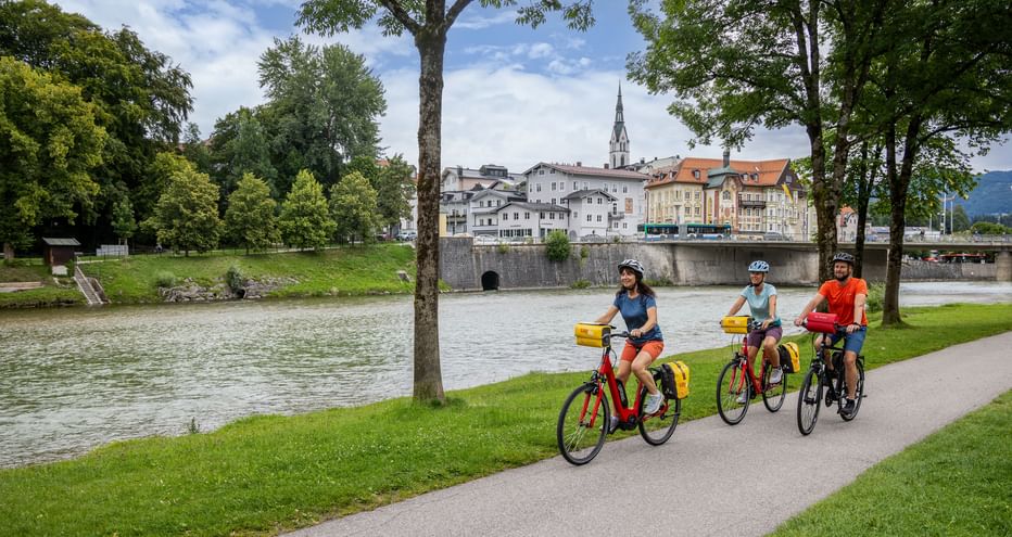 Three cyclists riding along the Isar River in Bad Tölz. Historic town buildings and church spire visible across the river under green trees.
