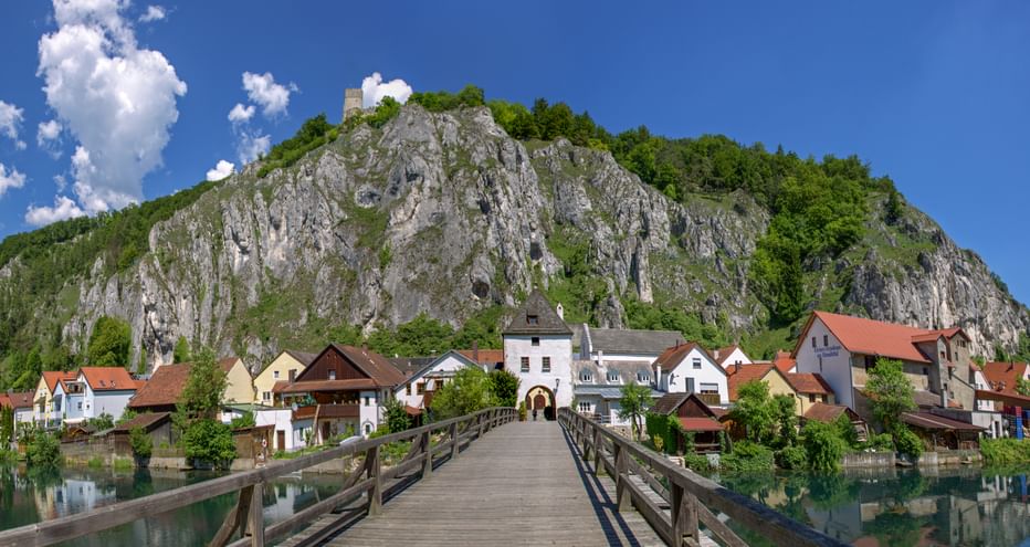 Eine Holzbrücke mit Blick auf Markt Essing im Altmühltal, im Hintergrund ein felsiger Hügel