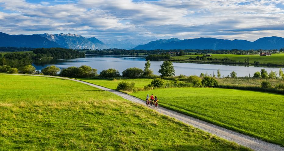 Three cyclists on beautiful cycle paths across meadows, with Lake Riegsee and the mountain panorama in the background